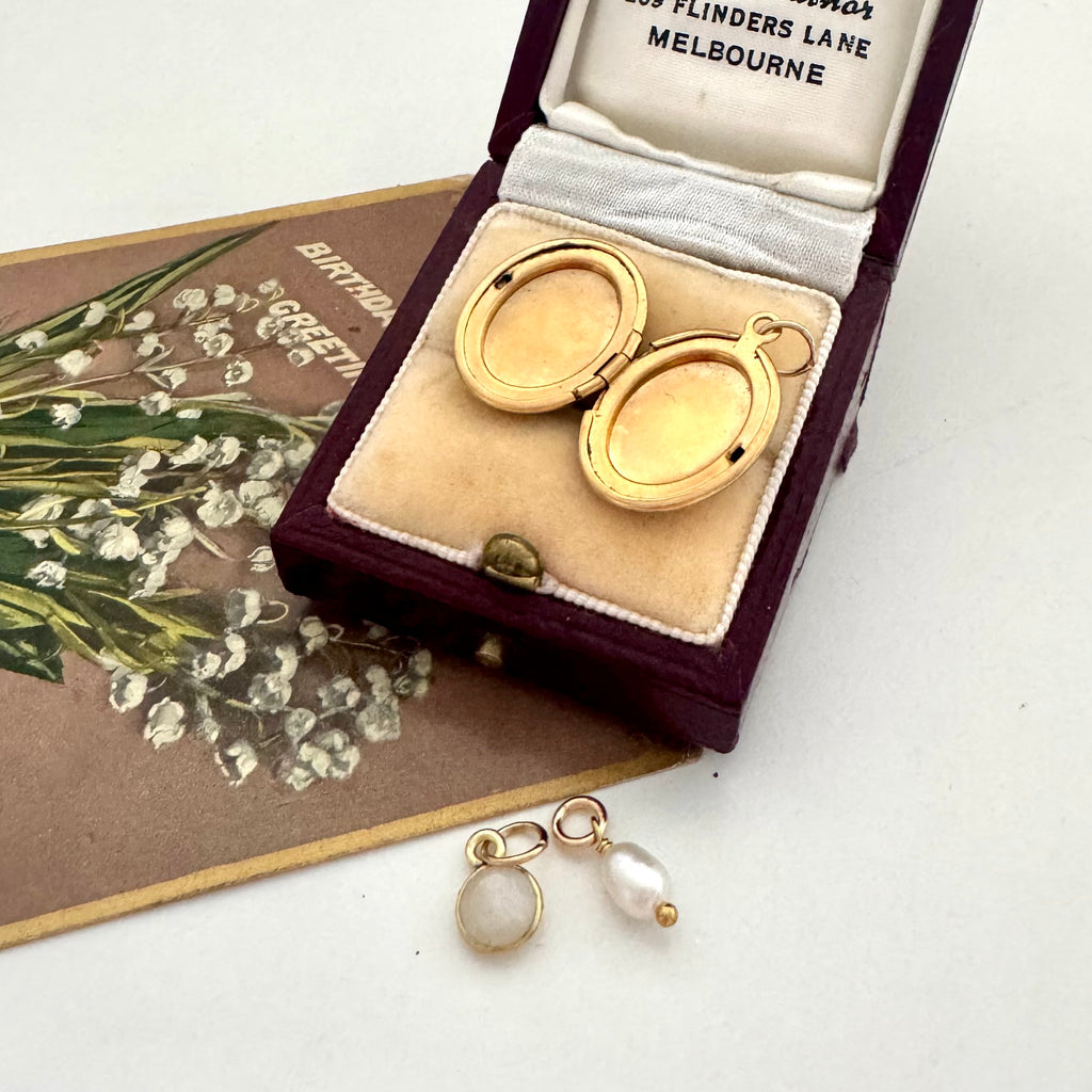 Gold locket with leaf design in a jewellery box and a pearl and moonstone charm on a white surface