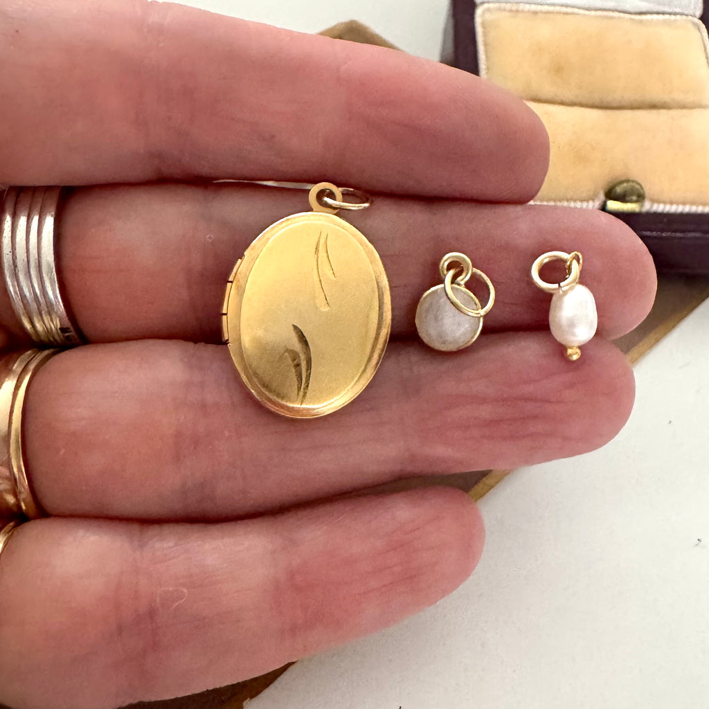 Gold locket and charms held between fingers with a jewelry box in the background.