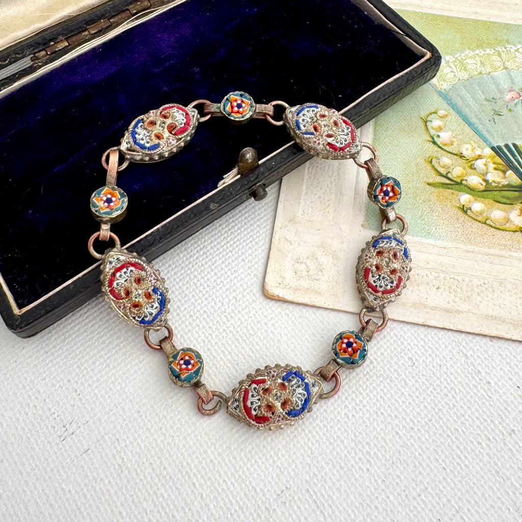 An ornate vintage micro mosaic bracelet with large oval and round enamel medallions in red, blue, white, and gold, displayed on a white textured surface next to an open dark velvet jewelry box and a decorative card.