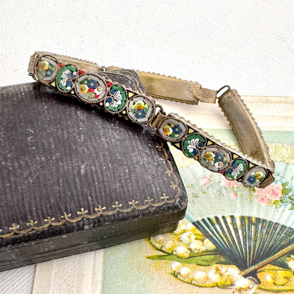 Avintage micro mosaic bracelet with a silver-toned metal frame decorated with colorful enamel medallions, resting on a dark vintage book next to a floral fan.