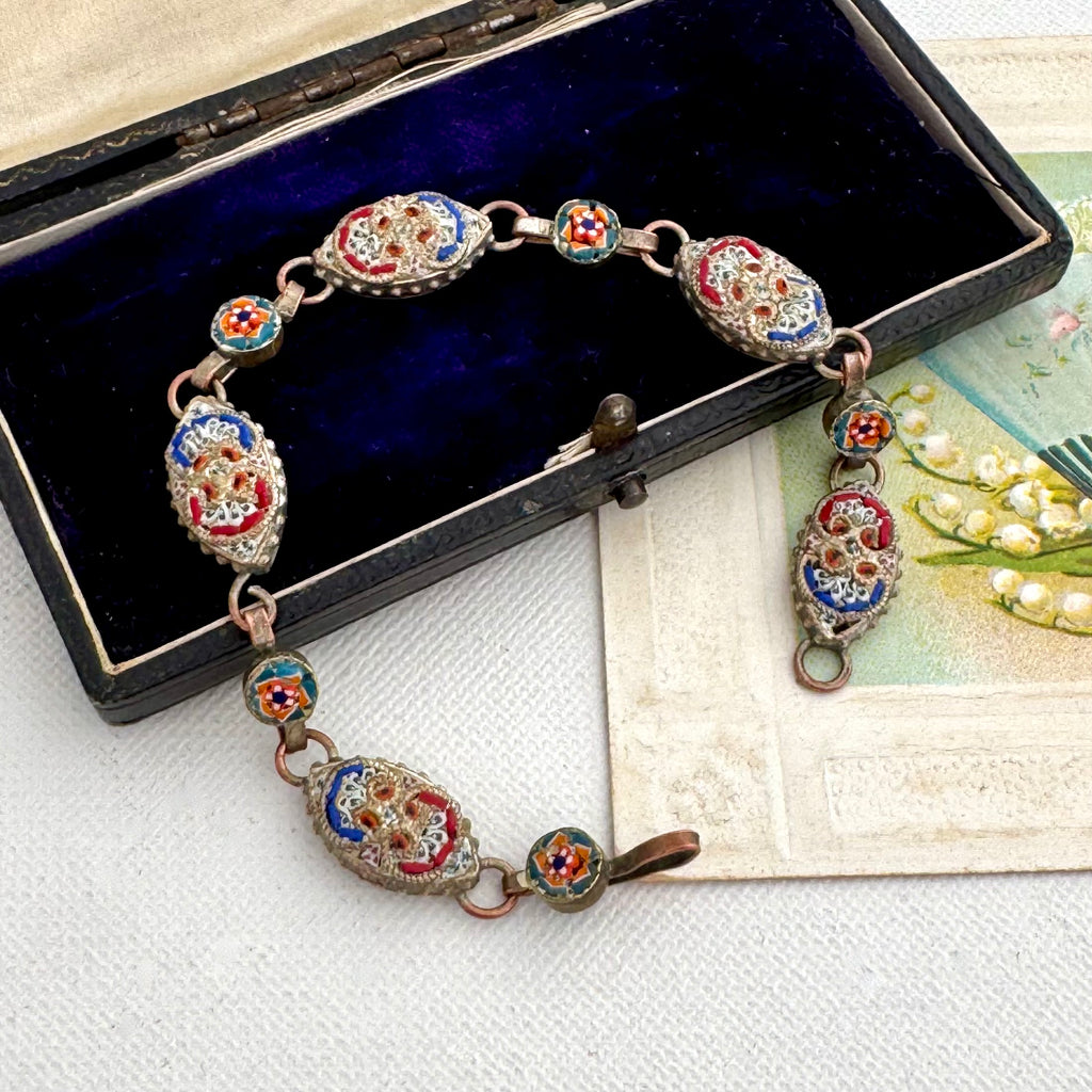 An ornate vintage-style bracelet with oval enamel medallions in red, blue, and yellow, displayed on a dark velvet jewelry box over a cream-colored card.