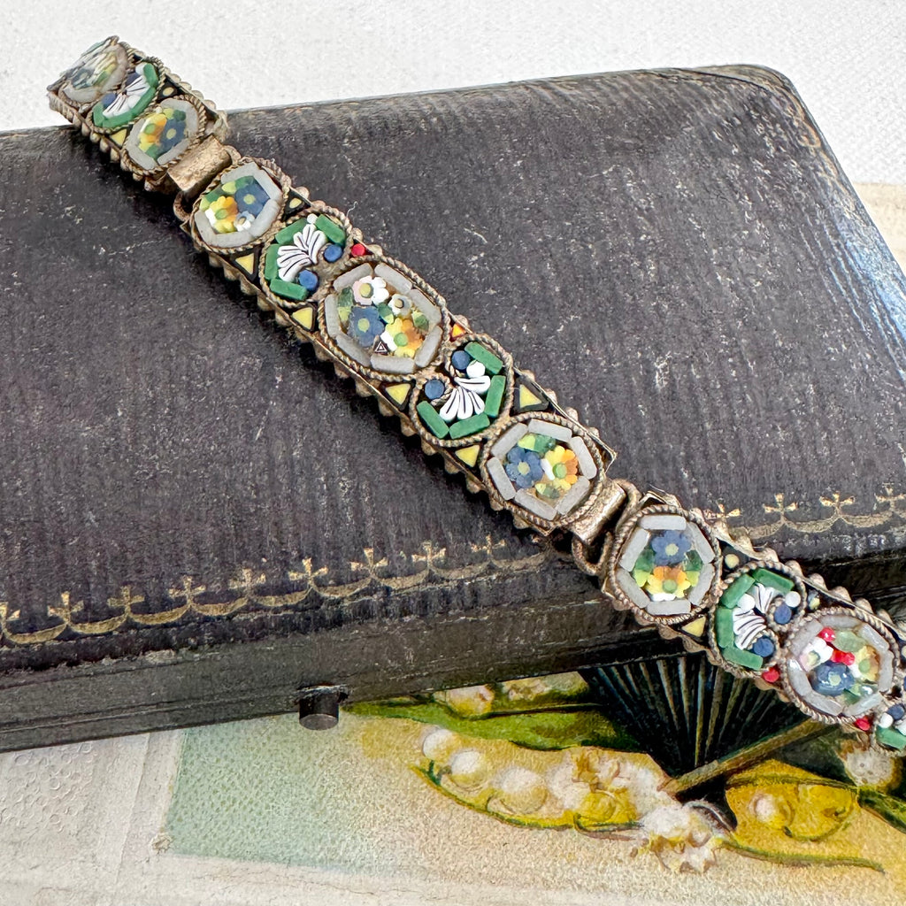 An ornate vintage micro mosaic bracelet with oval enamel inlays, resting diagonally on a dark textured leather case.