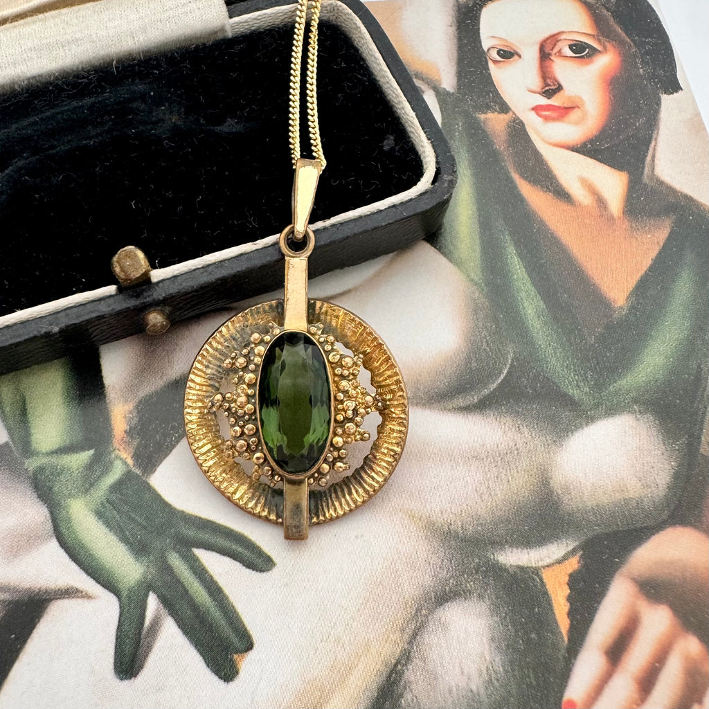 A vintage Andrea Daub gold pendant necklace featuring a large oval dark green gemstone surrounded by textured gold beads and a circular gold frame, resting on a vintage illustrated card.
