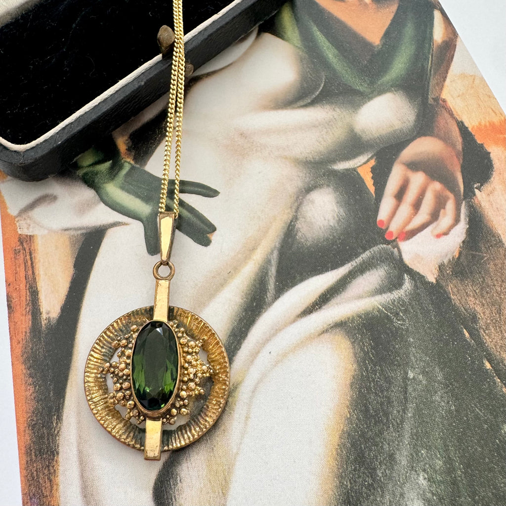 A vintage Andrea Daub gold pendant necklace featuring a large oval dark green gemstone surrounded by textured gold beads and a circular gold frame, resting on a vintage illustrated card.