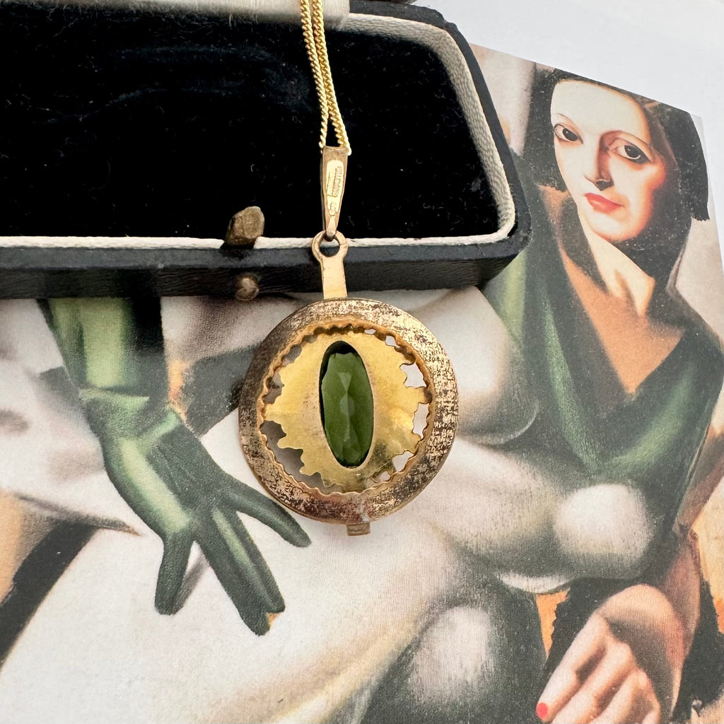Back of a vintage Andrea Daub gold pendant necklace featuring a large oval dark green gemstone surrounded by textured gold beads and a circular gold frame, resting on a vintage illustrated card.