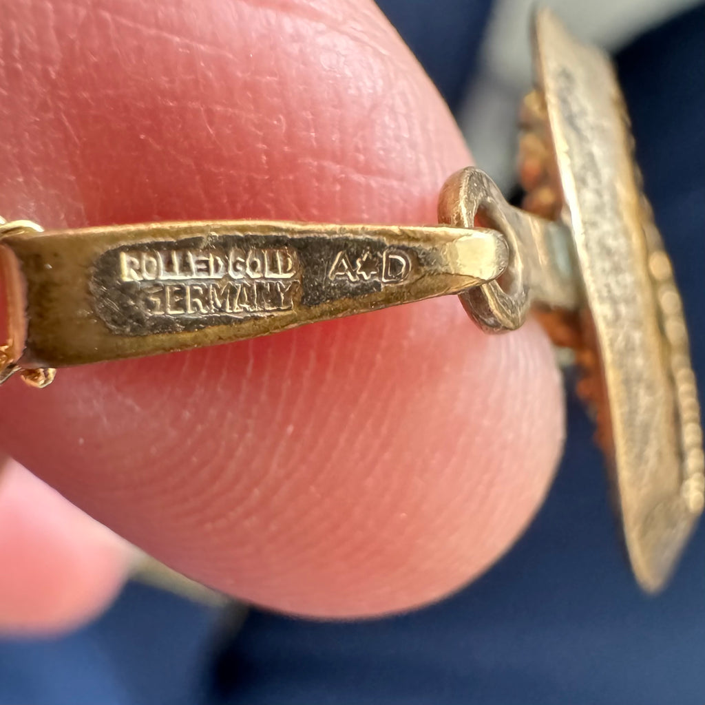 Close-up of a gold-toned piece of jewelry held between fingers, showing engraved hallmarks including 'ROLLED GOLD Germany' and 'A*D'.
