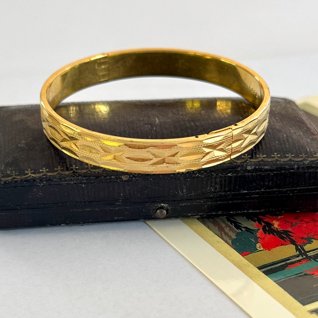 A gold bangle bracelet with an engraved geometric pattern, resting on a dark wooden box.