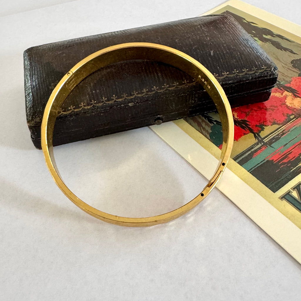 A gold-toned metal bangle bracelet displayed in front of a dark brown textured case, with a colorful printed card underneath on a light surface.