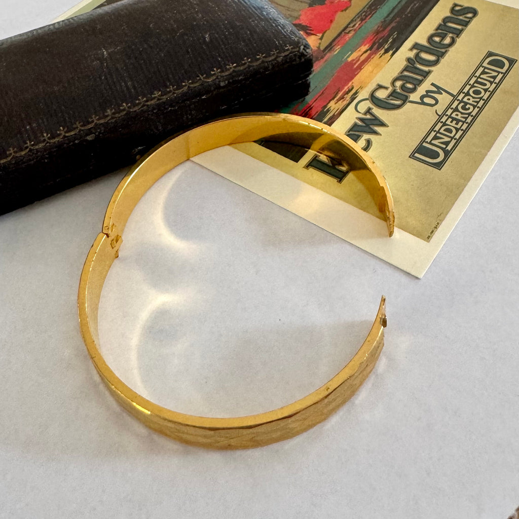 A polished gold-colored split cuff bracelet displayed on a light gray surface, with a dark textured case and a vintage New Carvards by Underground ticket in the background.
