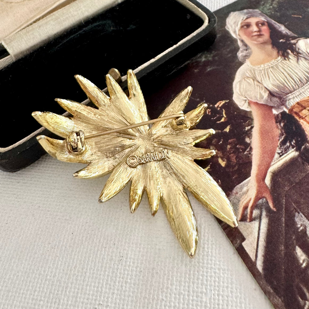 Back of signed Sarah Coventry brooch from 1965 Tropics Collection on a textured surface with an open jewelry box and vintage illustration.