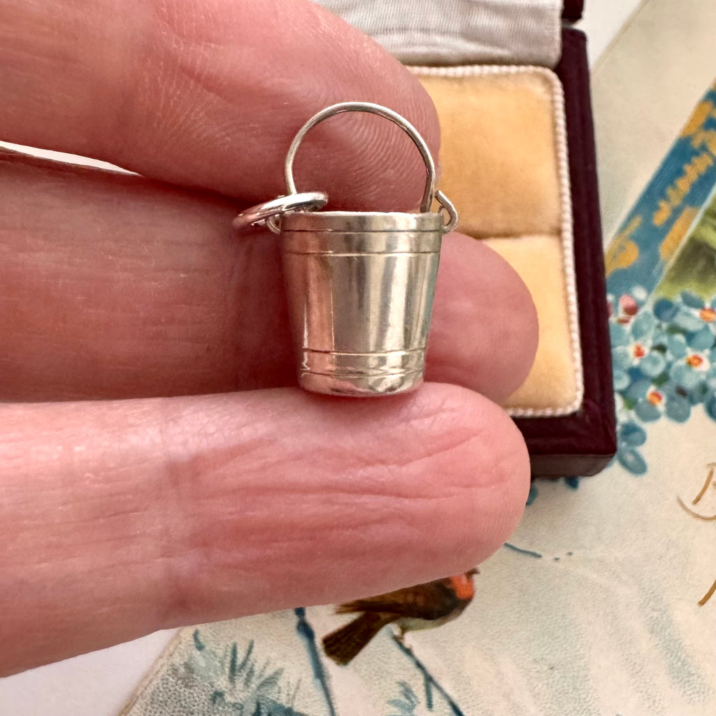 Small silver bucket pendant held between fingers with a decorative background