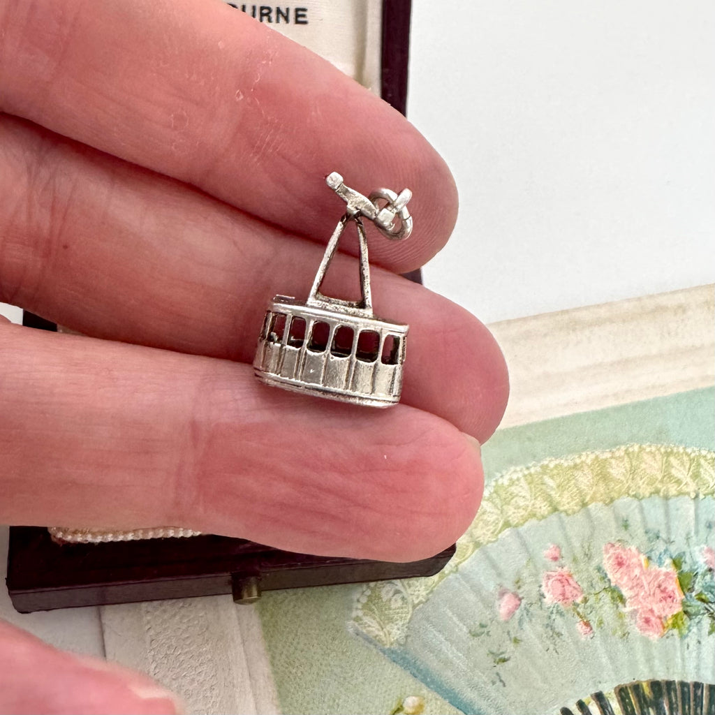 Vintage silver cable car charm held between fingers with a decorative fan in the background