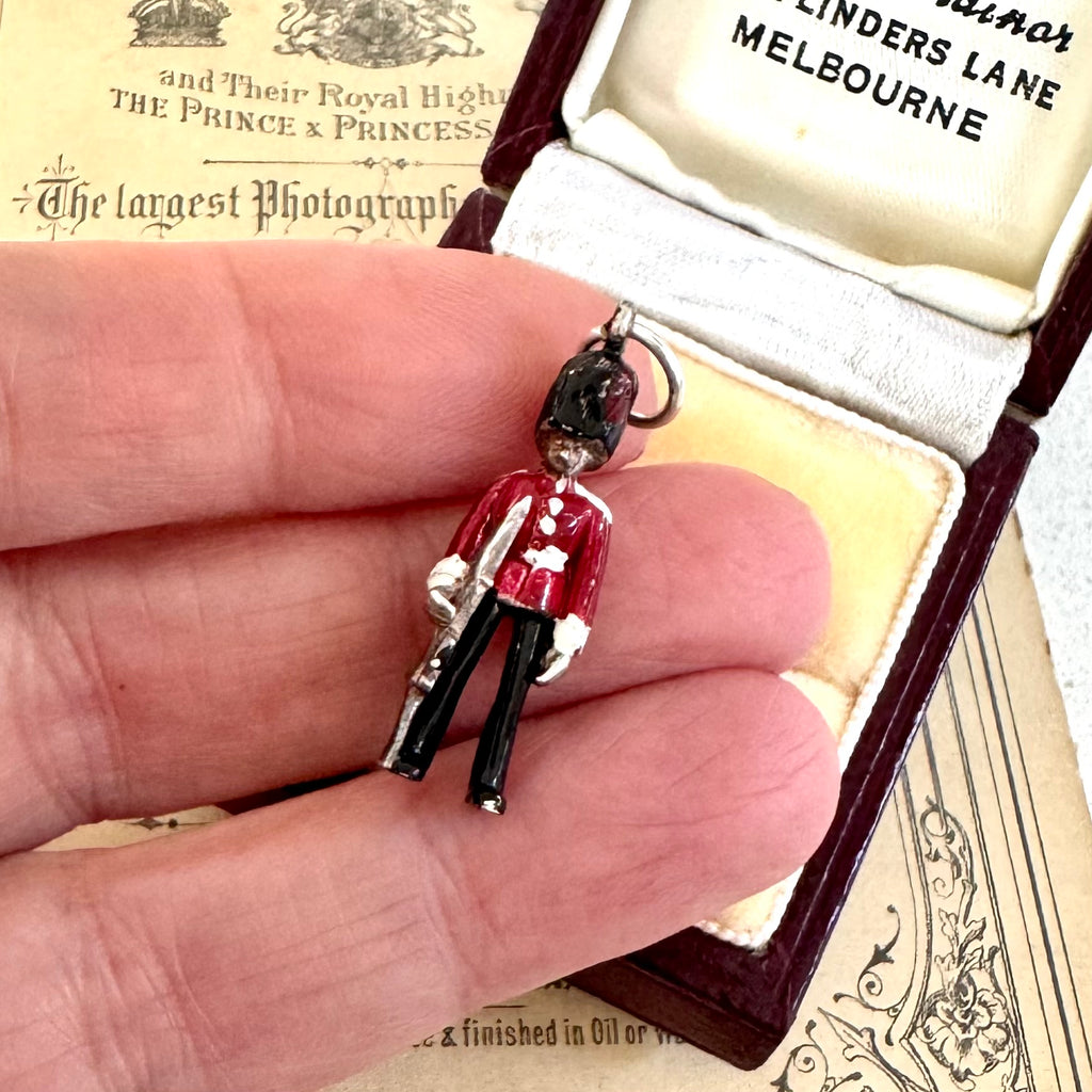 Vintage silver and enamel British Royal Foot Guard charm in someones hand and with an open jewelry box with vintage paper in the background.