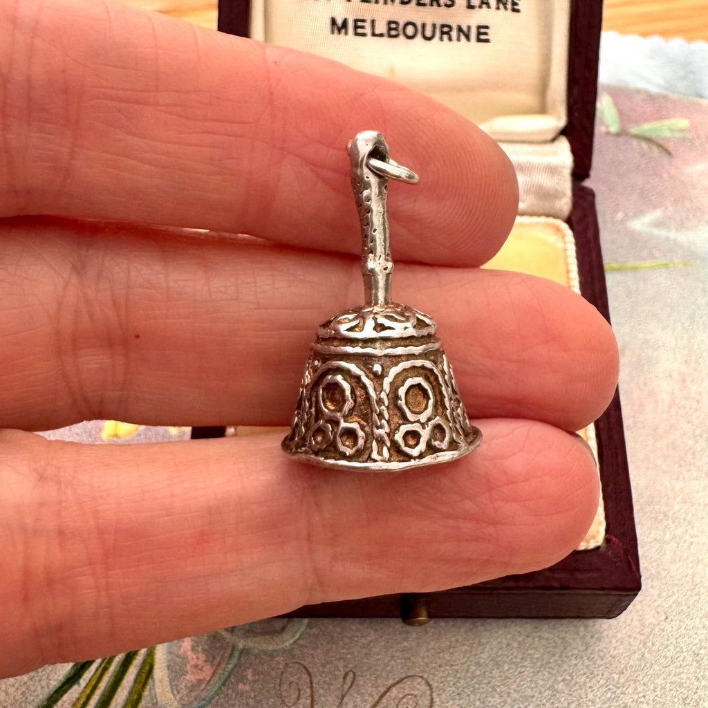 A vintage silver bell-shaped charm with ornate cutout detailing is held between a person's fingers, with an open jewelry box in the background.