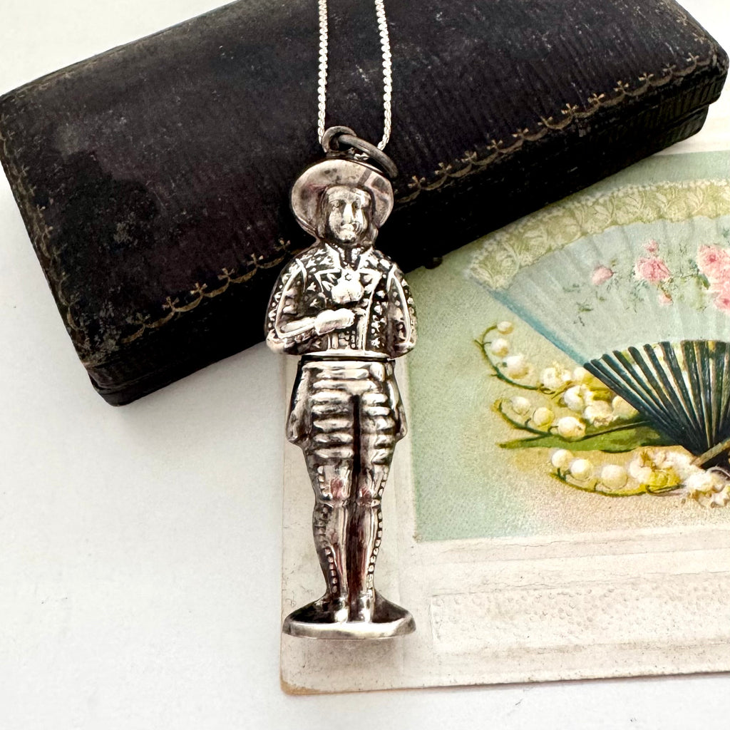 Vintage sterling silver needle case in the form of a boy holding a bunch of flowers. There is a vintage postcard and jewellery box in the background.