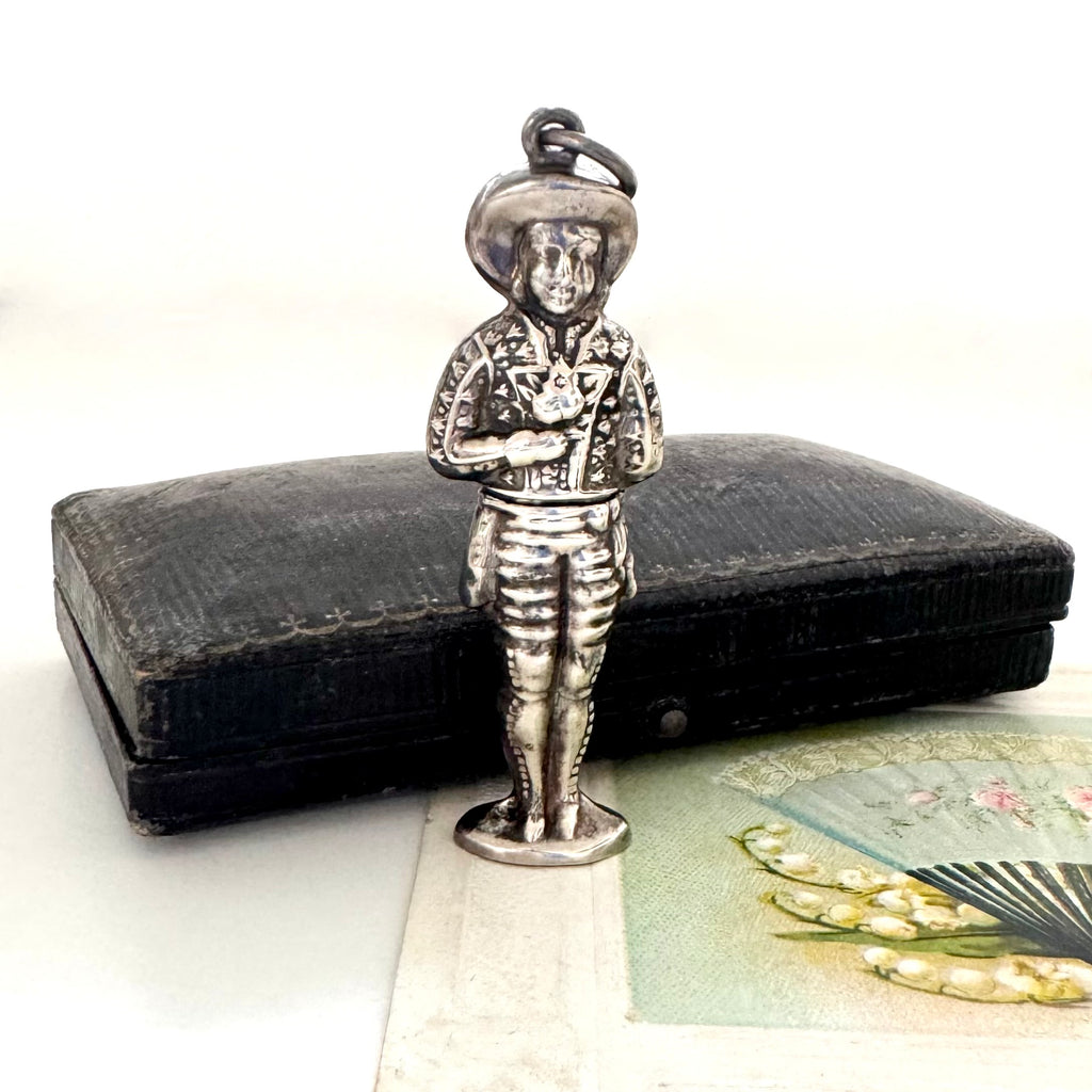 Vintage sterling silver needle case in the form of a boy holding a bunch of flowers. There is a vintage postcard and jewellery box in the background.