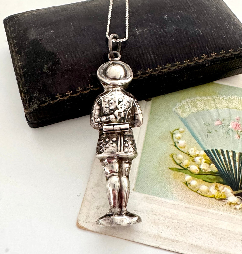 Vintage sterling silver needle case in the form of a boy holding a bunch of flowers. There is a vintage postcard and jewellery box in the background.