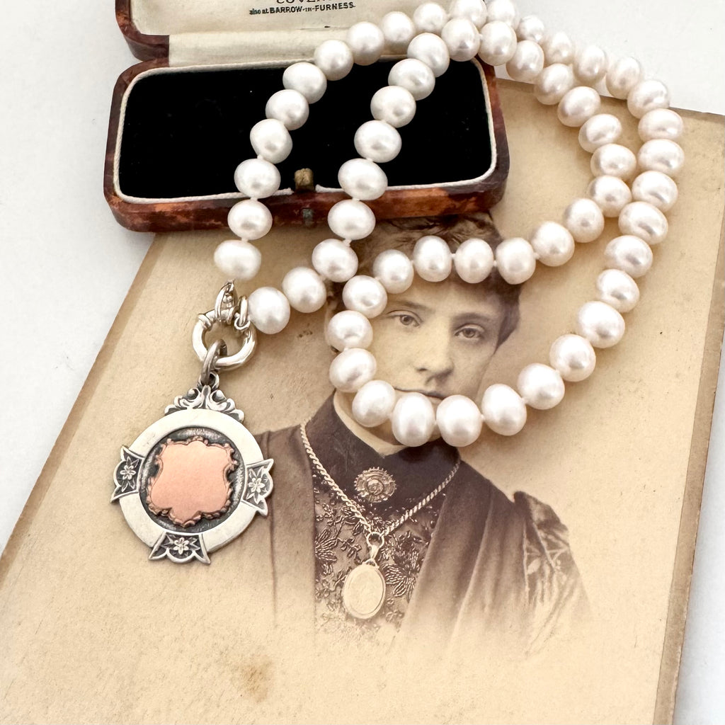 A strand of white pearl beads draped over an antique sepia-toned portrait print, with a sterling silver and rose gold watch fob and a vintage jewelry box in the background.