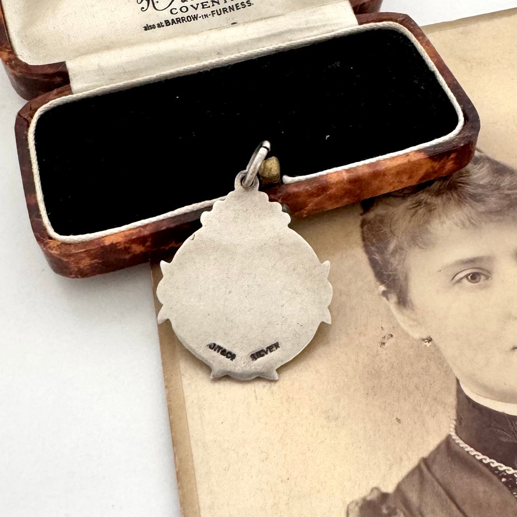 A vintage sterling silver watch fob pendant with a scalloped border and small engraved text, displayed in front of an open vintage wooden jewelry box on top of an old sepia portrait photograph.