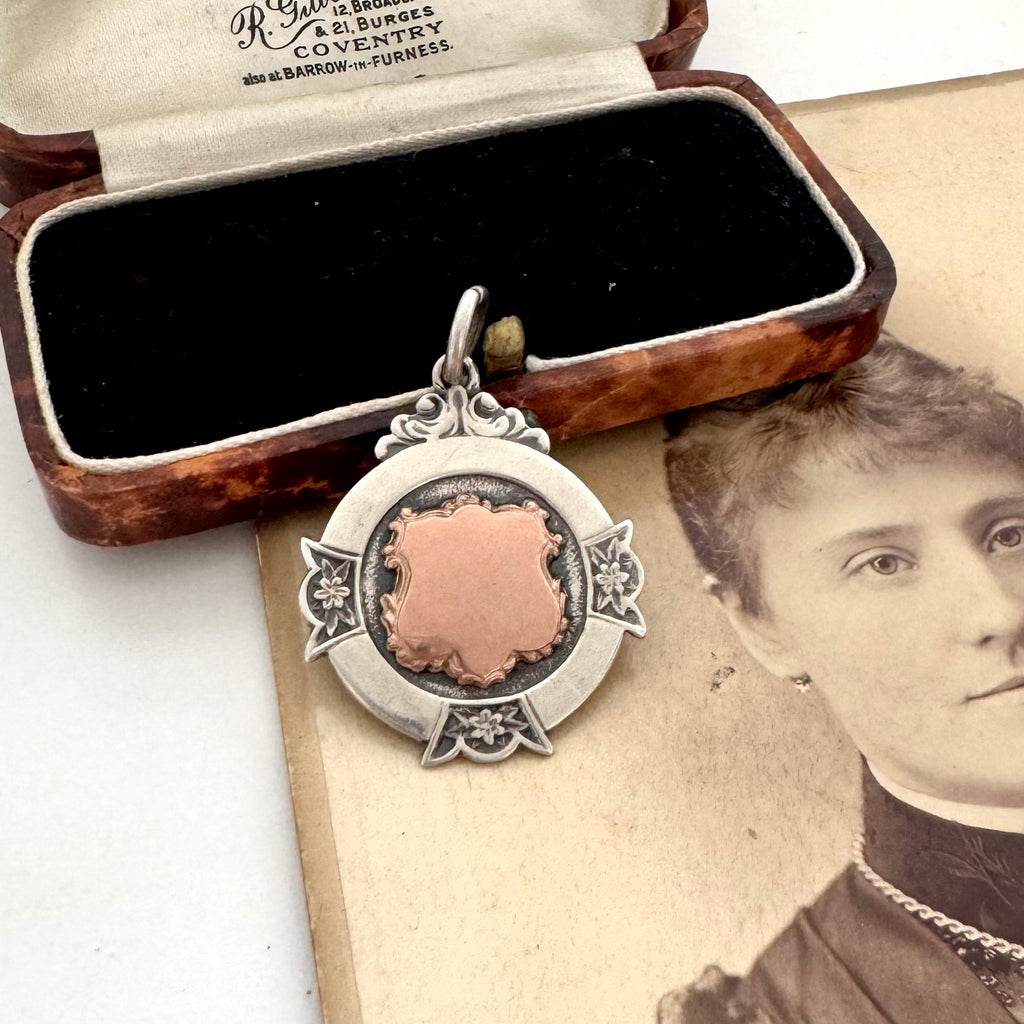 A vintage sterling silver watch fob pendant with a rose-gold shield-shaped center, displayed next to its original wooden and velvet box on top of an antique sepia portrait photograph.