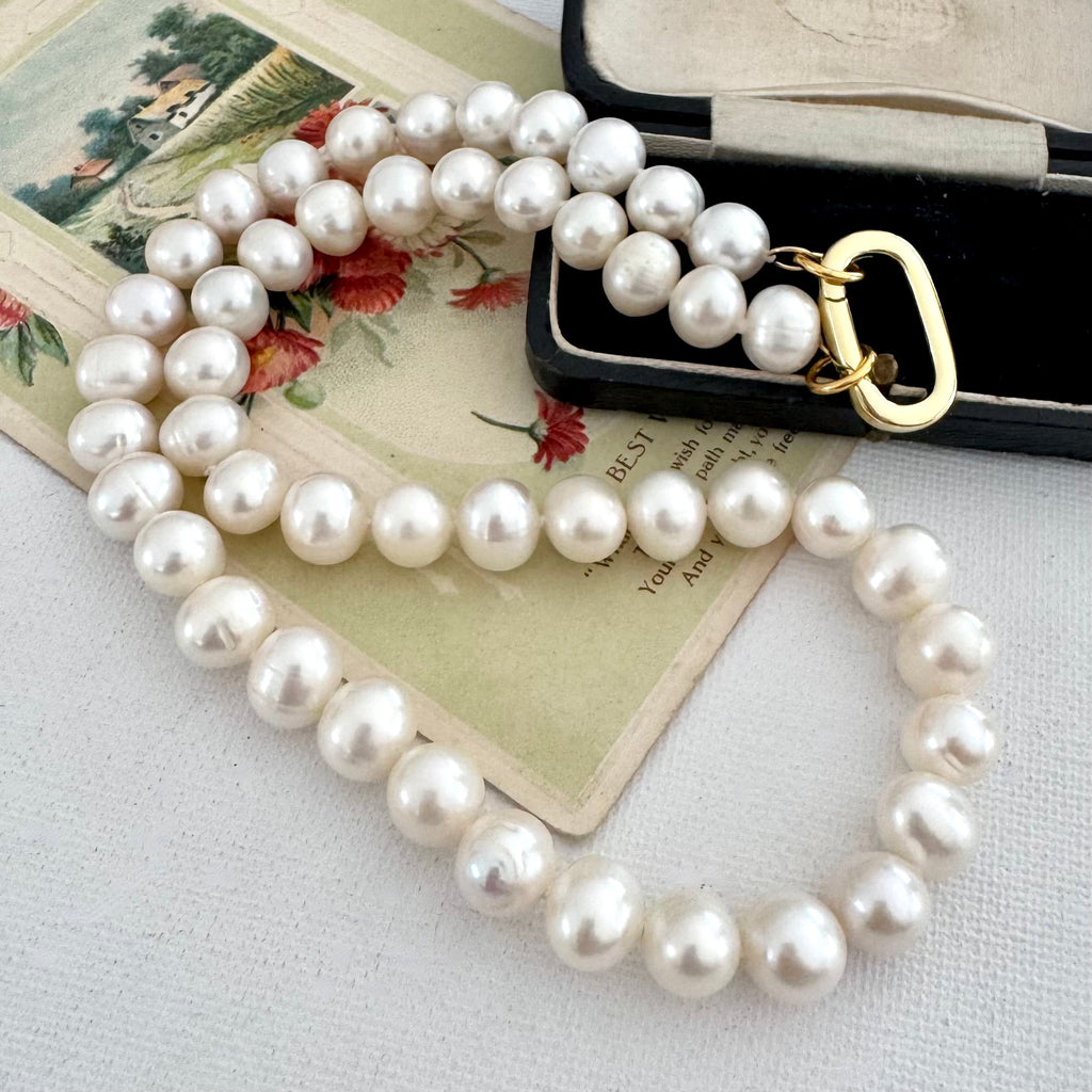 A strand of white freshwater pearls with a gold clasp, draped over a vintage floral card and resting on a black jewelry box.