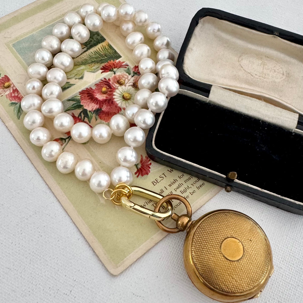 A vintage-style pearl strand necklace with a gold clasp displayed next to an open black velvet jewelry box and a gold pocket watch, arranged on an antique floral card over a white fabric background.