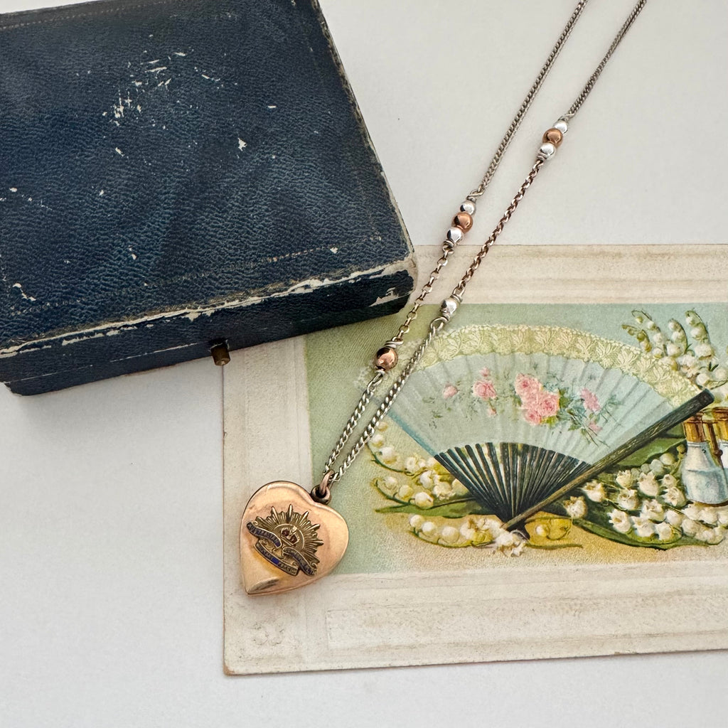 WW2 ACMF heart-shaped pendant sweetheart locket on a chain against an old postcards with fan illustration