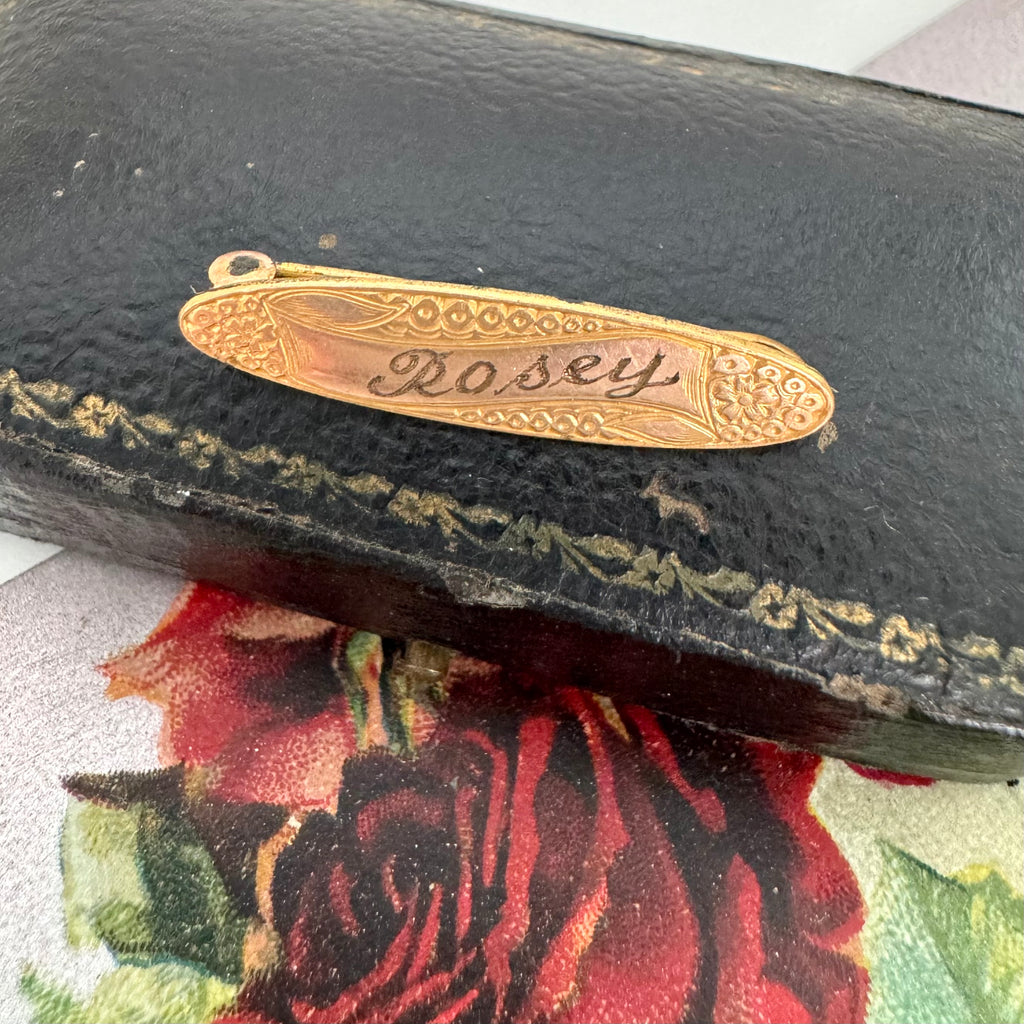Gold antique brooch with 'Rosey' engraved on it, attached to a black leather case.