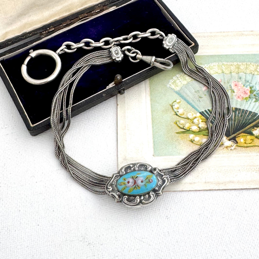 Antique French silver albertina chain with an oval turquoise and floral enamel centerpiece, displayed on a white surface with an open black presentation box and a decorative card in the background.