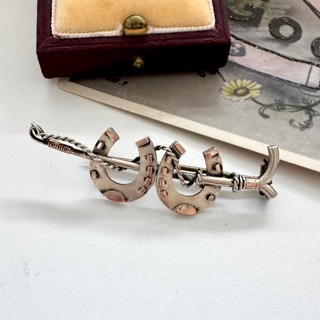 Antique silver horseshoe and riding crop brooch on a white surface with a jewelry box in the background.