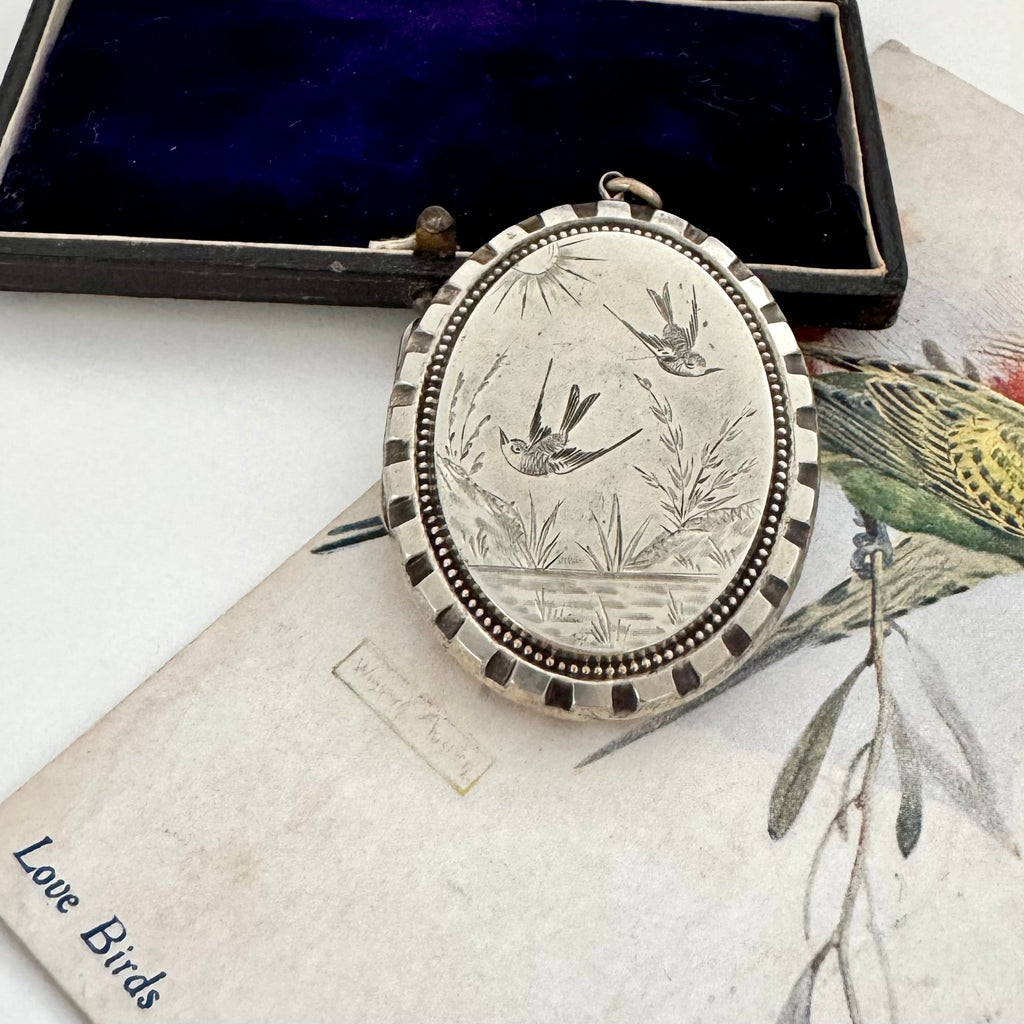 Silver Victorian swallow locket with engraved design on a vintage box and botanical illustration