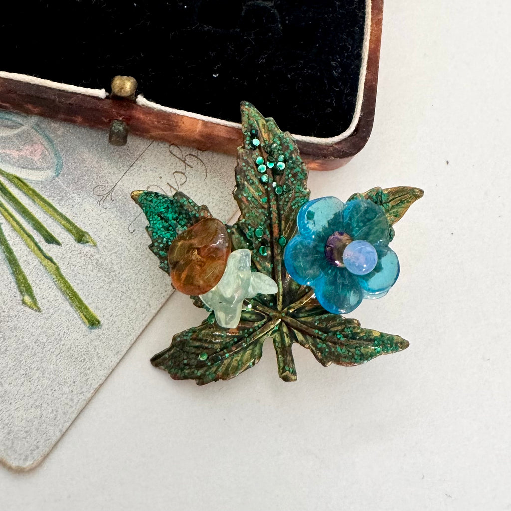 A vintage-style brooch featuring a green glitter-leaf design with a blue flower, a light green stone, and a brown stone, displayed on a light surface next to a black velvet case.