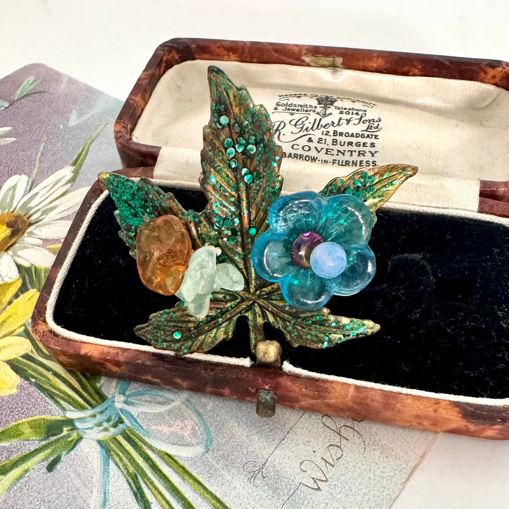 A vintage brooch shaped like a leaf with turquoise and amber stones, displayed inside an open antique jewelry box with a black velvet lining.