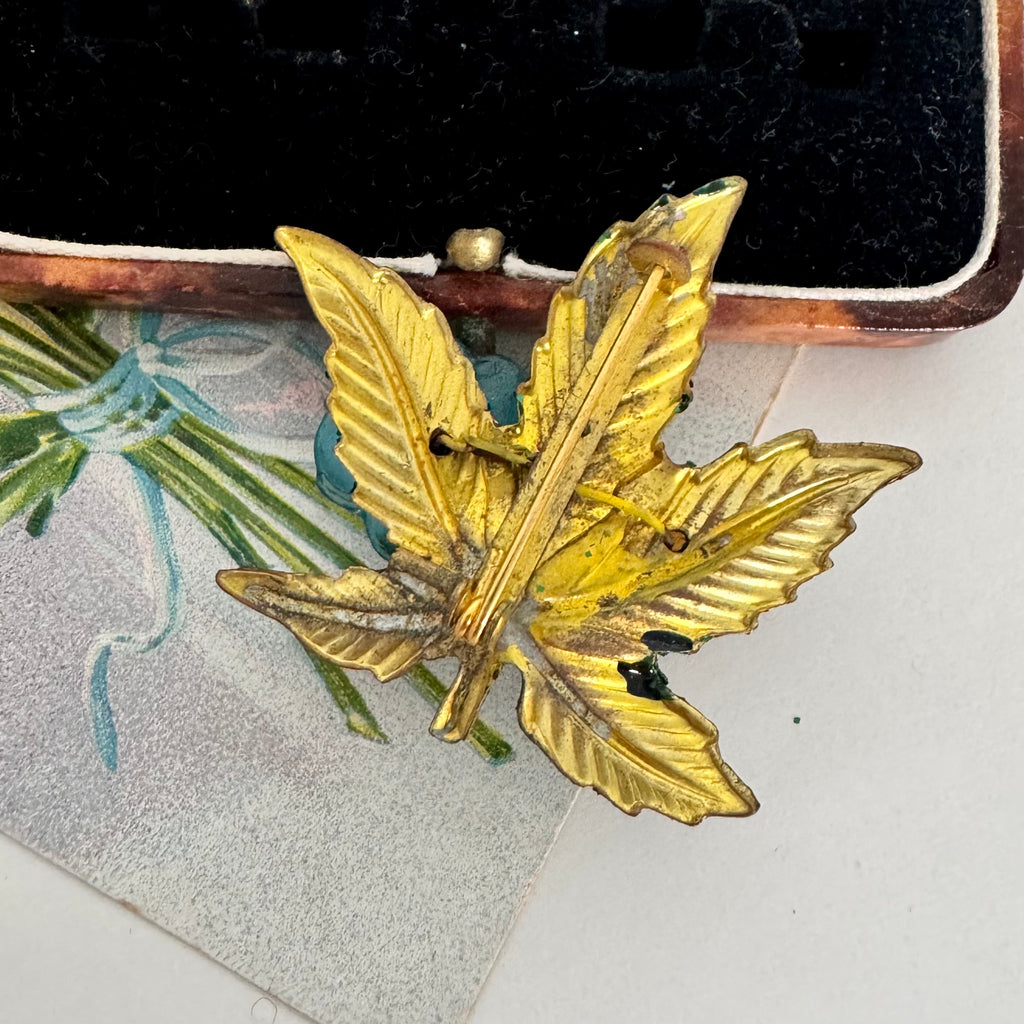 A gold-toned metal leaf-shaped brooch with a textured, vintage finish, displayed on a decorative card over a black velvet jewelry box.