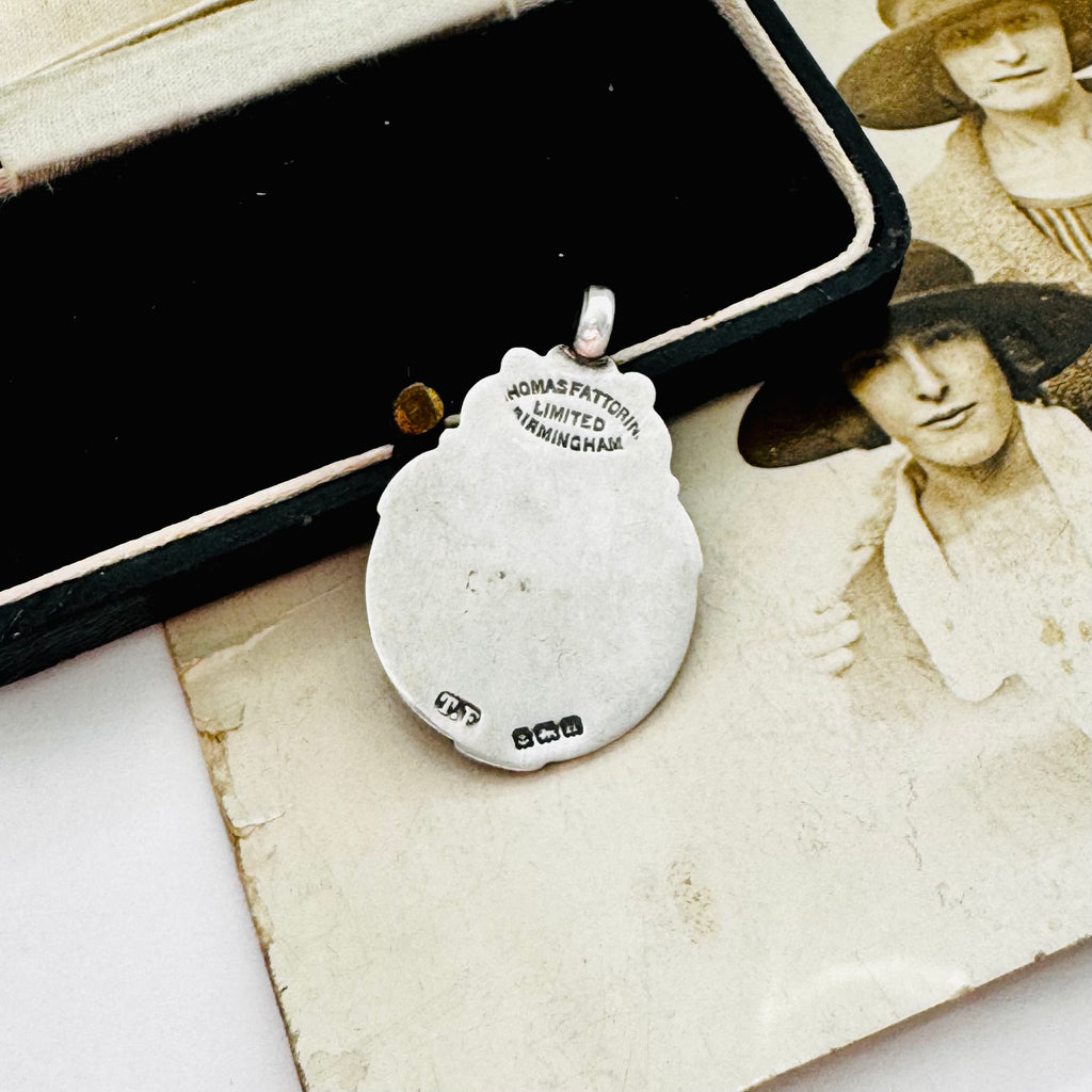 Back of vintage 1932 Fattorini sterling silver 1932 watch fob pendant with ornate detailing, displayed on top of an antique sepia-toned photograph and a black jewelry box.