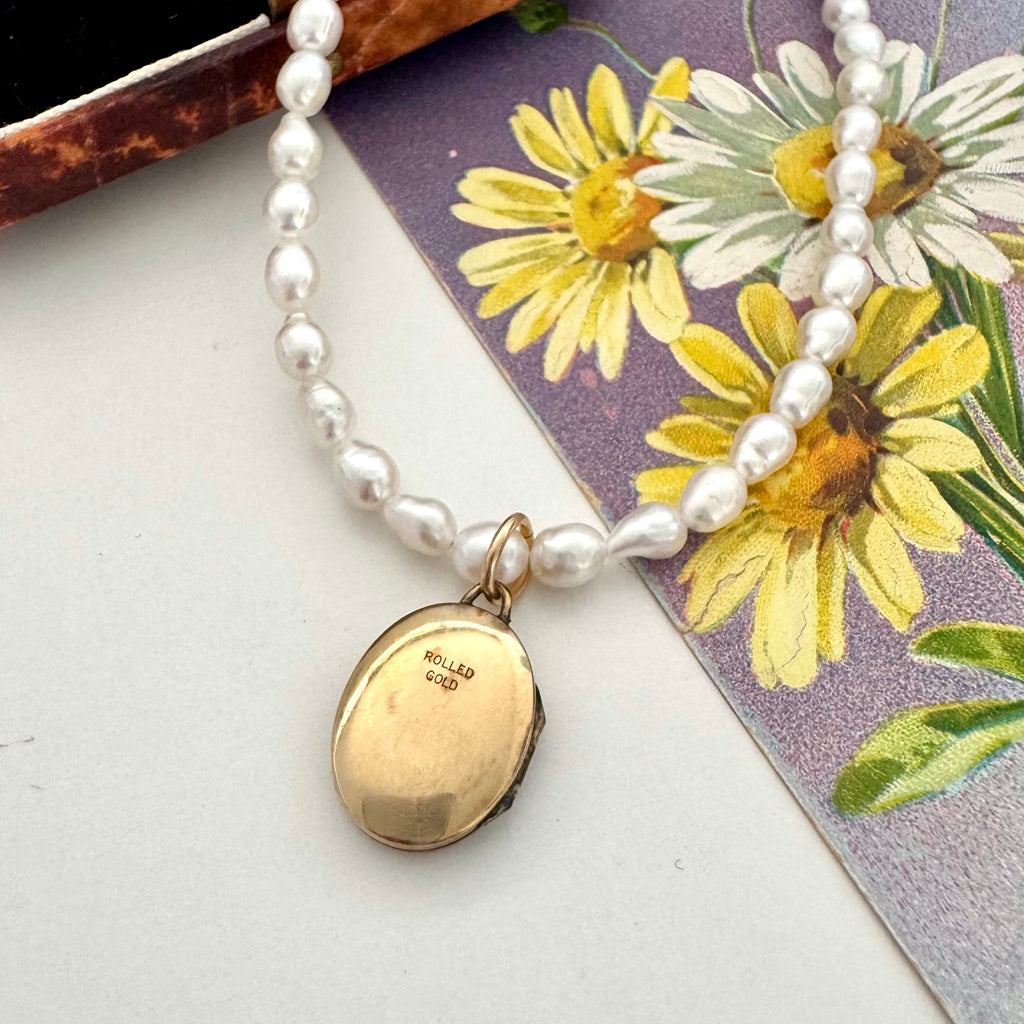 A pearl strand necklace with a gold oval locket pendant, displayed on a floral card over a light surface. The locket is engraved with 'Rolled GOLD'.
