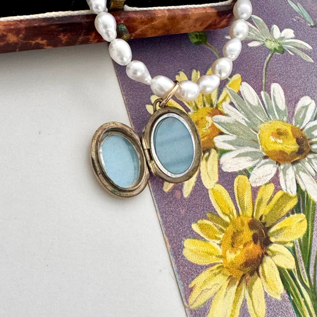 A close-up of a pearl beaded necklace with an open oval locket pendant featuring light blue inlay, resting on a floral card with daisies and a purple background.