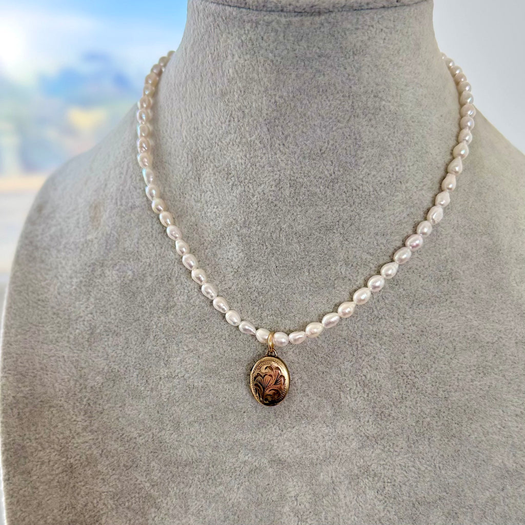 A necklace of irregular white pearls with a small oval vintage rolled gold locket, displayed on a gray felt jewelry bust.
