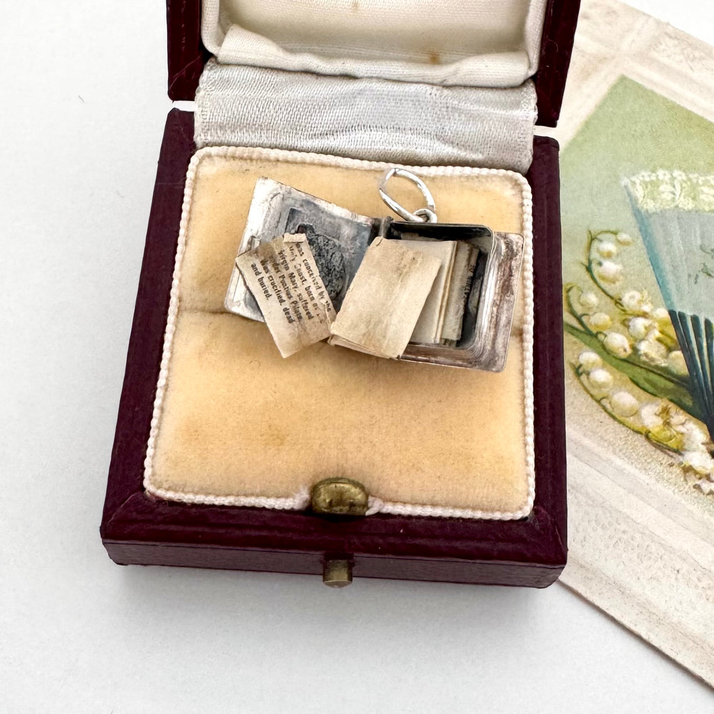 A miniature silver book-shaped pendant with a silver ring clasp, displayed inside an open vintage burgundy velvet jewelry box with a cream lining. The box is placed on a white surface next to a decorative card featuring lily of the valley flowers.