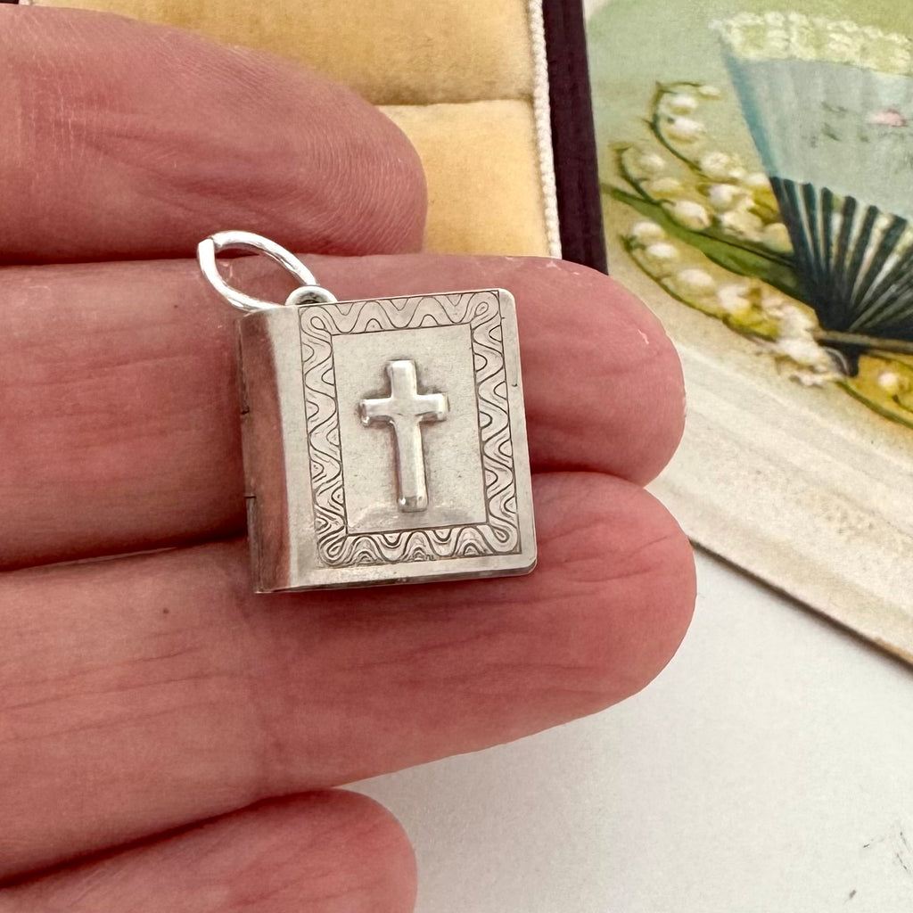 A small silver book-shaped pendant with a cross on the front, held between fingers to show its size. The pendant has engraved decorative borders and a silver bail at the top.