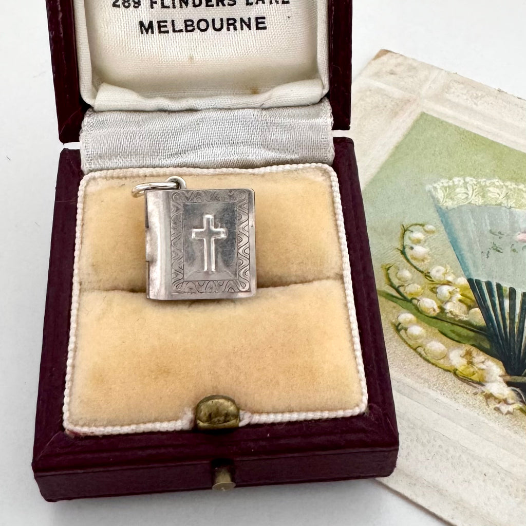 A silver cross pendant or charm displayed inside an open vintage presentation box with cream velvet lining and a dark burgundy exterior. The box lid is open, showing printed text inside, and a decorative card is partially visible beside it.
