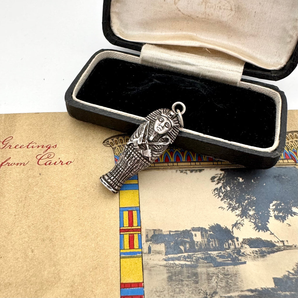 Silver pendant of a pharaoh in an open jewelry box with a scenic postcard underneath.