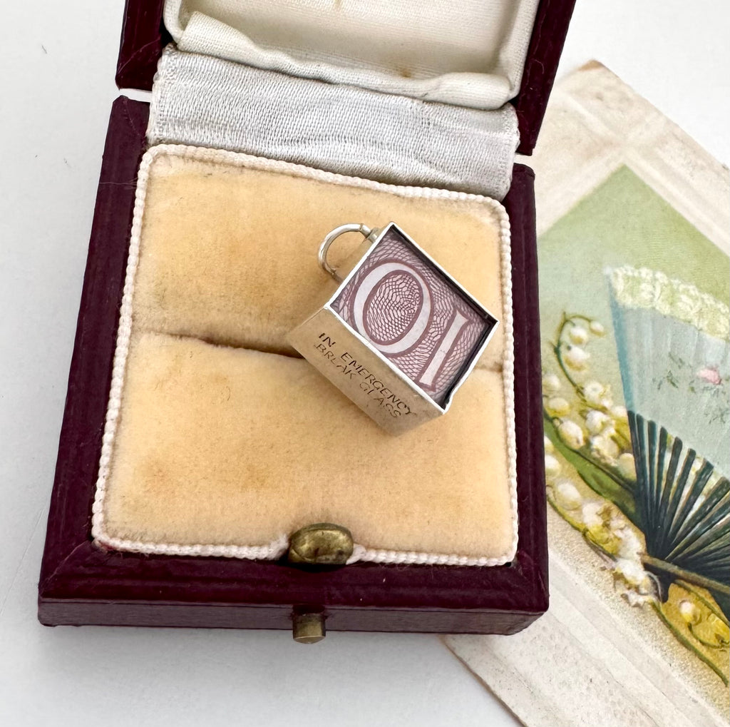 A vintage emergency money silver charm t, displayed inside an open dark wood jewelry box with beige velvet lining.