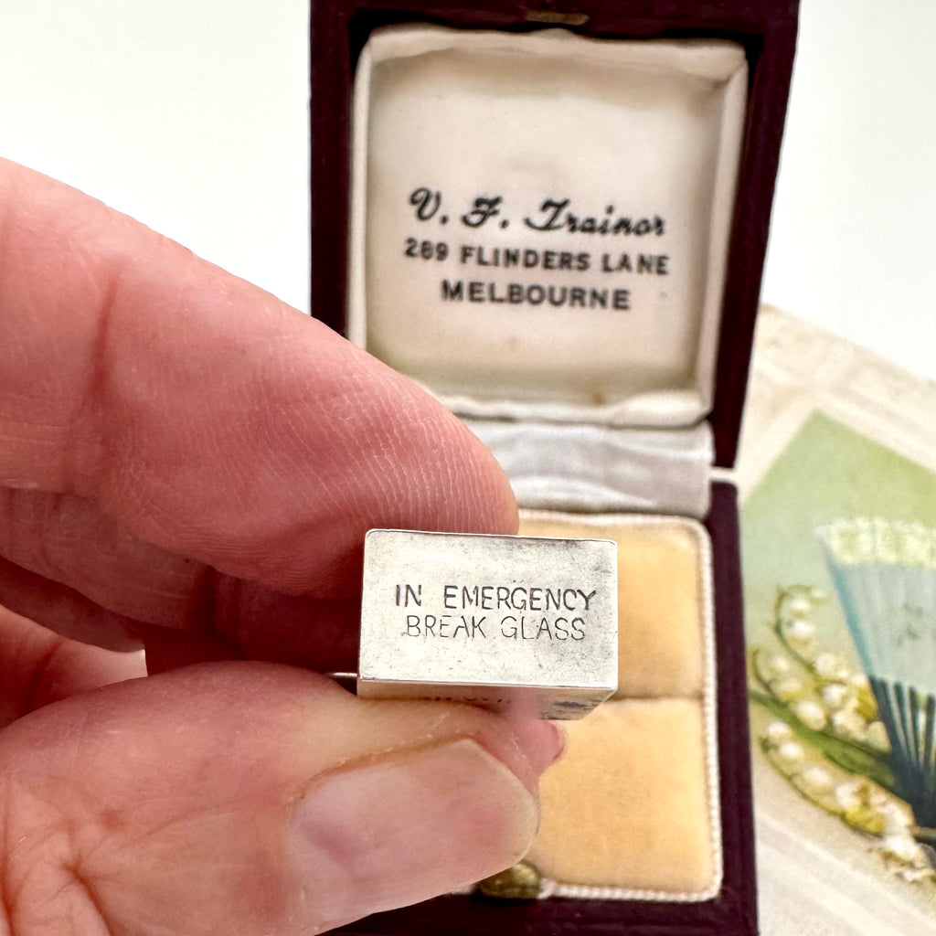 A hand holds a small silver emergency money charm engraved with 'IN EMERGENCY BREAK GLASS' in front of an open vintage dark red presentation box with cream lining and printed address text.