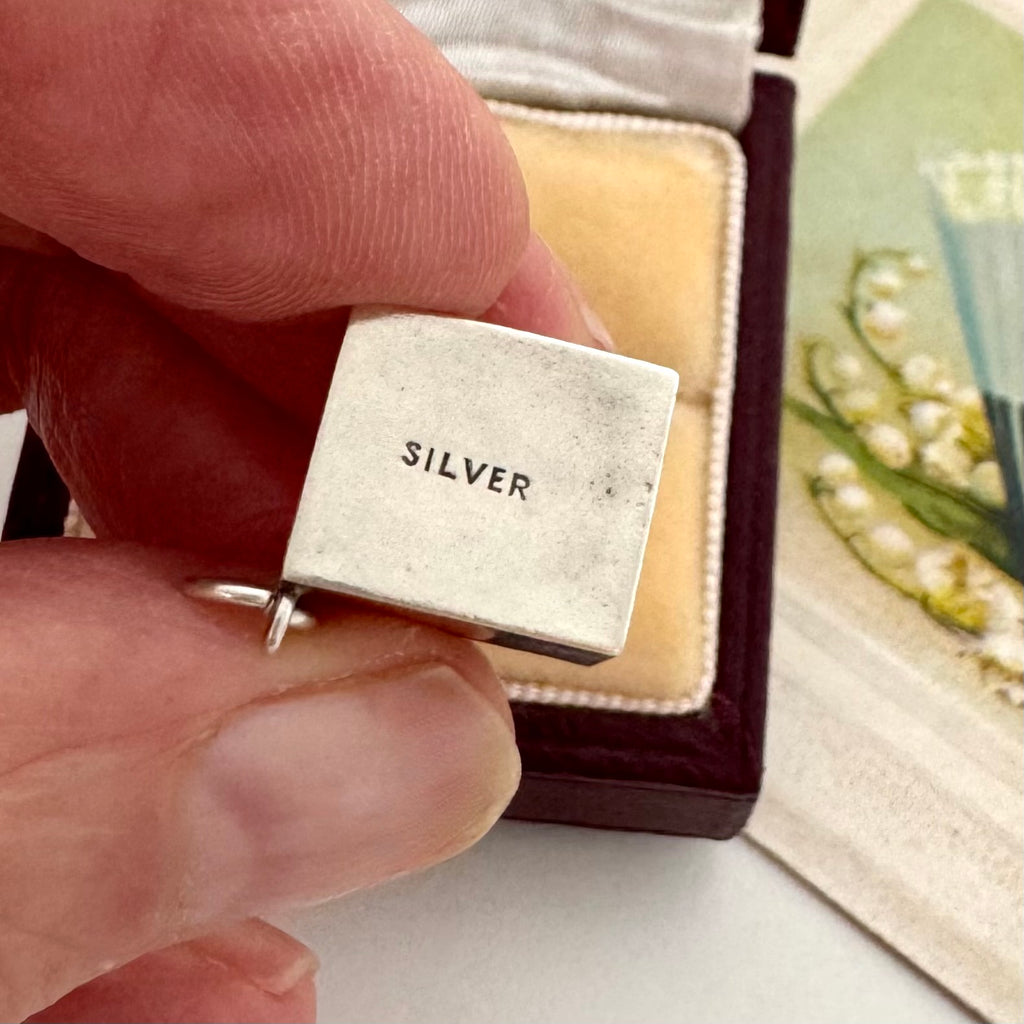 A close-up of a hand holding a small silver charm engraved with the word 'SILVER', positioned in front of an open jewelry box.