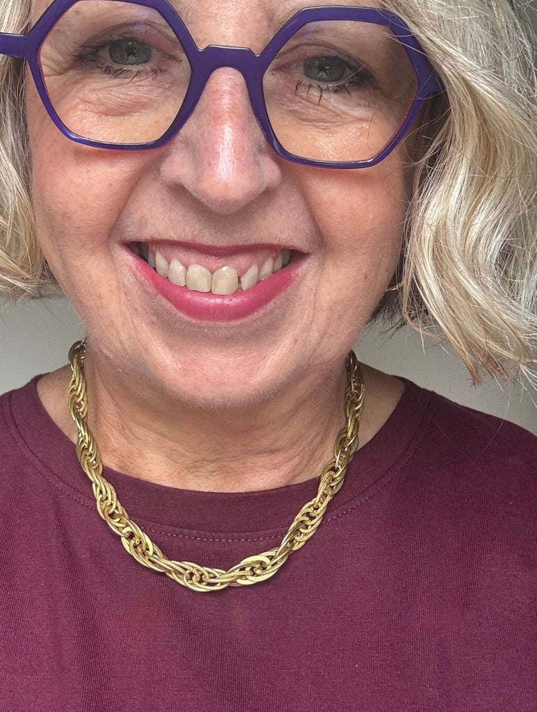 A close-up of a smiling woman with gray hair wearing purple geometric eyeglasses and a chunky gold chain necklace, paired with a burgundy top.