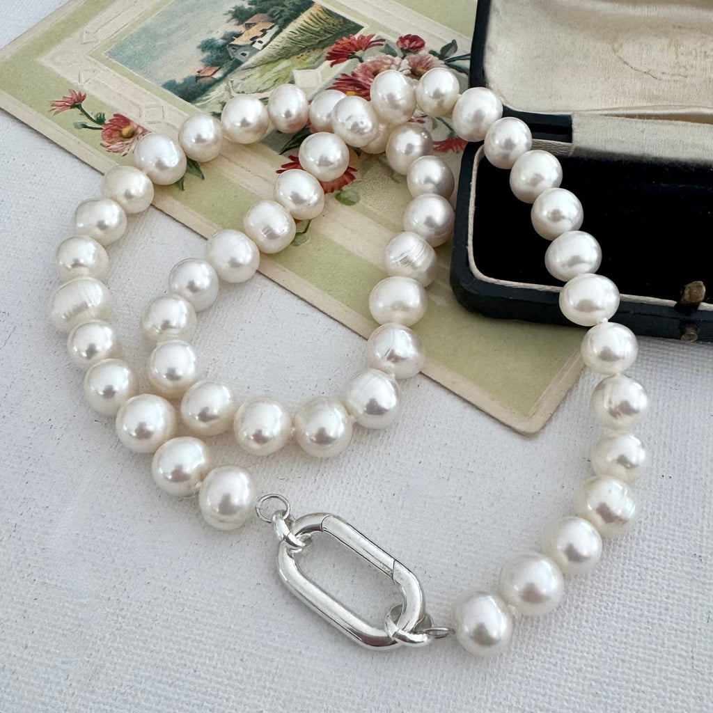 A strand of lustrous white pearls arranged in loops with a polished silver-toned oval clasp, photographed on a light fabric surface with a vintage floral card and a black jewelry box as props.