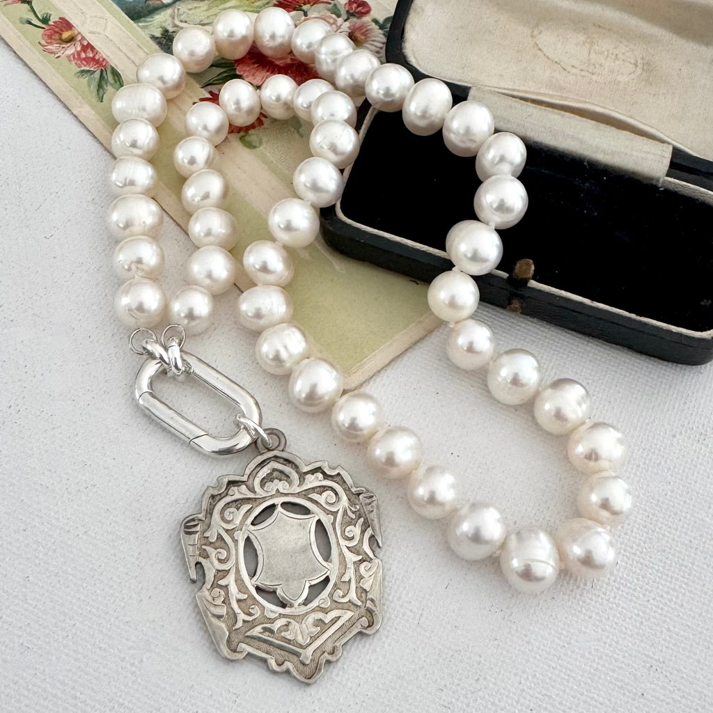 A strand of white freshwater pearls draped over a vintage floral card, with a silver-toned decorative pendant clasp and a black presentation box in the background.