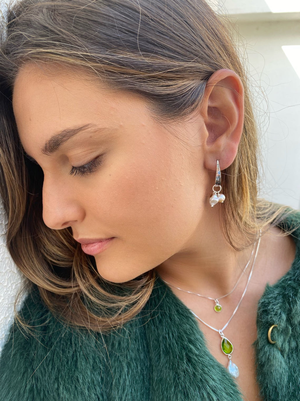 Girl with dark hair with head resting against a wall. She is wearing Leoni & Vonk jewellery and a green fur jacket.