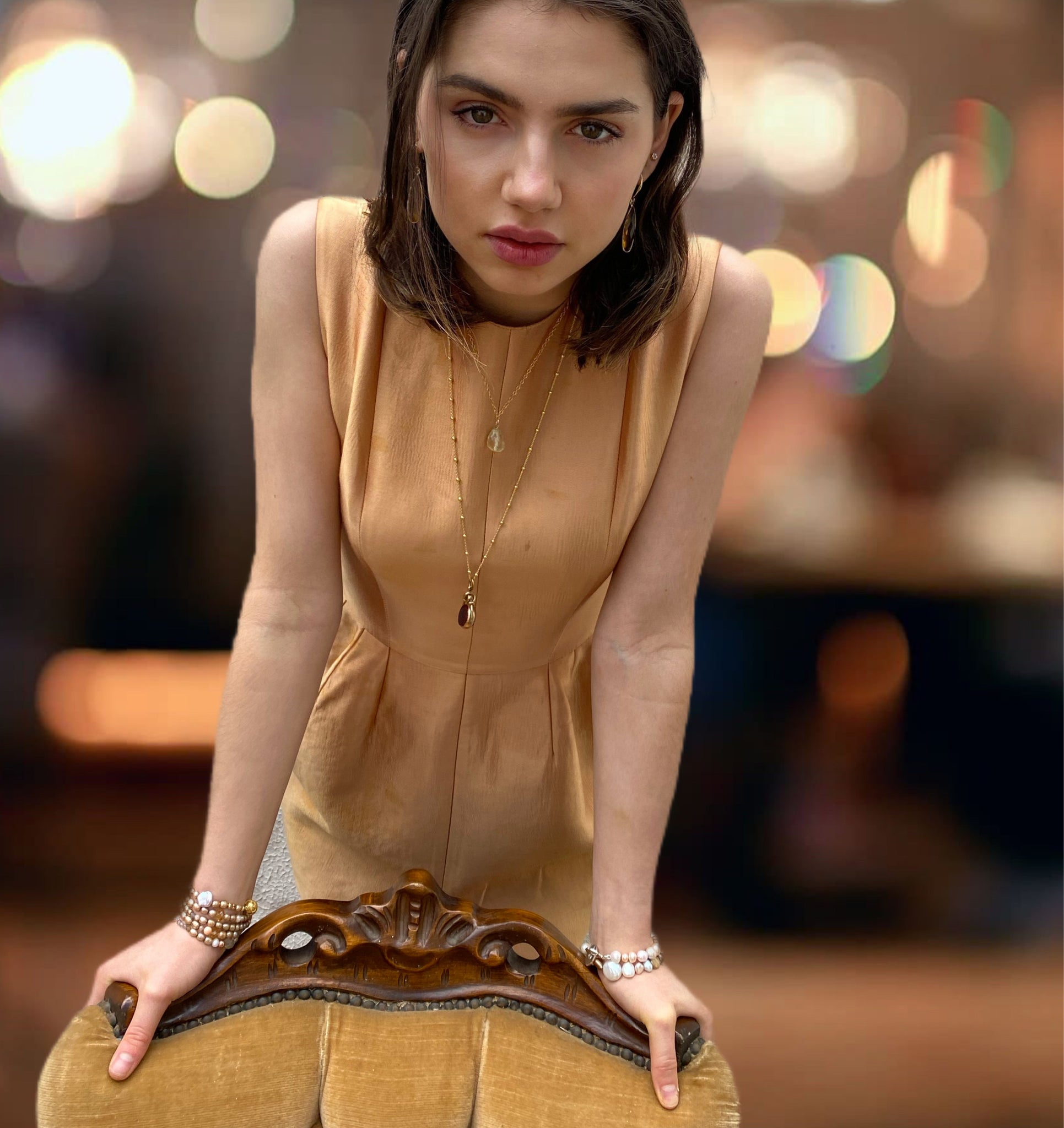 Dark haired model wearing a gold Ellery dress and Leoni 7 Vonk jewellery. She is leaning over a gold velvet chair